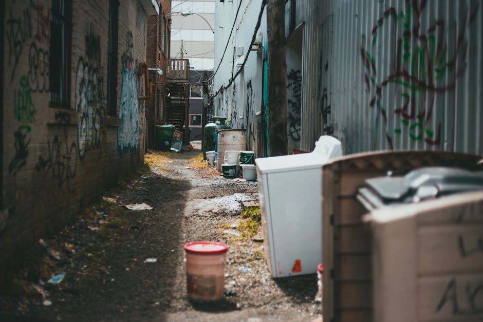 The image depicts a narrow alleyway between two buildings, with graffiti-covered brick and metal walls on either side. The ground is uneven, composed of dirt and gravel, and appears slightly muddy. On the right side, there are large, white household appliances, including a refrigerator and washing machine, stacked and partially obscured, suggesting removal or disposal. Further along the alley, several green garbage bins and a white plastic container are positioned against the walls, some with lids open or placed on their sides. Scattered trash and debris can be seen along the path, including a red and white plastic bucket in the foreground. The alley is dimly lit, with natural light coming from the far end, where a set of external wooden stairs leads to an upper level of a residential building. This scene illustrates a typical backstreet area where private rubbish collection or waste removal services, such as those provided by rubbishremovalkingstonuponthames.com, could be necessary to clear accumulated waste and problematic clutter from narrow and hard-to-access urban spaces.