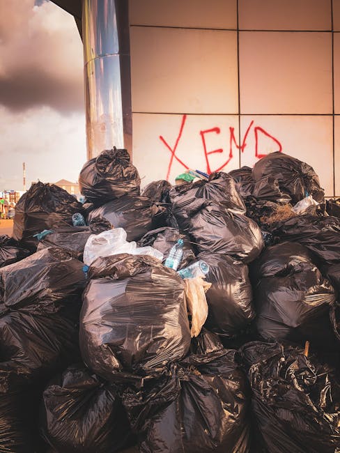 A narrow urban alleyway flanked by brick and concrete buildings, filled with a large, overflowing recycling or rubbish collection trolley made of woven synthetic material in a weathered dark grey color, positioned centrally in the foreground. Behind the trolley, there is a pile of mixed waste, including cardboard boxes, black plastic bags, and other refuse, stacked haphazardly against a brick wall with some debris scattered on the wet and slightly stained asphalt ground. To the right, a metal fence with vertical bars runs along the building, while to the left, there are graffiti tags on the brick surface. The environment appears dimly lit, possibly during late afternoon or early evening, with leafless tree branches visible overhead and in the background. The scene suggests a location where independent rubbish collection or on-site clearance activities might take place, reflecting the context of alternative waste handling solutions outside of standard municipal refuse services. This image is associated with private rubbish removal services on Norbiton narrow streets, as featured on rubbishremovalkingstonuponthames.com.