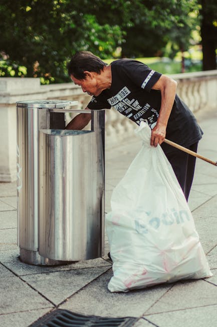 A middle-aged man with short dark hair, wearing a black T-shirt with white text and black pants, is seen collecting litter from a publicly accessible outdoor waste bin. The man is bending forward slightly, using their right hand to reach into a round, polished stainless steel bin to dispose of waste, while holding a large white plastic bag filled with rubbish in their left hand. The bag appears to contain general waste and is being held upright, supported at the bottom by the man's hand. The scene is set on a paved pathway or sidewalk with a low decorative stone wall adjacent to lush green trees and foliage in the background, suggesting a park or urban outdoor space. The surrounding environment is well-lit with natural daylight, and the overall scene reflects an act of personal waste disposal, which may be related to alternative rubbish management options, aligning with the theme of non-local-authority waste collection services, as supported by the context of rubbish removal services like those offered by rubbishremovalkingstonuponthames.com.