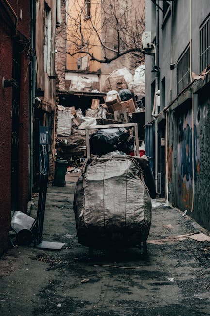 A large pile of mixed household waste and rubbish stored around a grey metal bin in an urban parking area. The rubbish includes cardboard boxes, plastic bags, paper, packaging, and assorted household items. Several black and green rubbish bags are scattered on the pavement, some leaning against the bin and others on the ground. The bin is filled with clutter, with some waste overflowing and resting on top. To the left, a parked silver car is visible, with a yellow license plate, behind a metal railing. In the background, there is a commercial building with blue scaffolding on the façade, storefront signage, and a small tree on the pavement, indicating a city or town centre environment. The scene suggests an overflowing rubbish collection point, highlighting the need for private waste management services like those offered by rubbishremovalkingstonuponthames.com as an alternative to standard council collection methods.