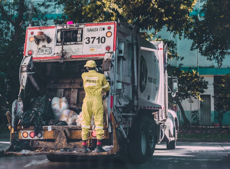 A waste collection truck fitted with a large rear hopper is actively engaged in rubbish removal, with a sanitation worker dressed in a yellow safety suit and helmet operating the lifting mechanism. The truck's exterior body is painted in a light grey colour with visible dirt and grime, indicating regular use. Several white and black garbage sacks, along with miscellaneous waste, are piled inside the open hopper, ready for disposal or transfer. The vehicle is positioned on an urban street, with some green-leaved trees and a modern building in the background, providing natural daylight that illuminates the scene. The surrounding environment suggests an area designated for waste collection, possibly on a municipal or private site. This scene represents the routine activity undertaken by waste management companies, such as rubbishremovalkingstonuponthames.com, which specialise in private waste disposal services. The truck's design and waste materials emphasize the importance of efficient rubbish removal and alternative waste handling outside traditional council services, supporting a professional approach to rubbish collection and debris clearance.