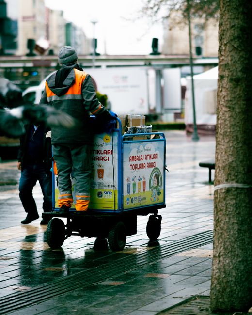 A person wearing a dark jacket with orange reflective stripes, a grey beanie, and matching orange and grey trousers, is pushing a blue wheeled cart loaded with large recycling bins and containers on a wet, paved outdoor street. The bins have printed labels and images related to waste collection, including text indicating they are for container waste and recycling. The scene is set on a city sidewalk with a tree trunk visible on the right, and blurred background elements include buildings, a metal railing, and an urban environment suggesting an area where private waste disposal or on-site clearance services, such as those provided by rubbishremovalkingstonuponthames.com, might be involved in managing waste outside of standard municipal collection services. The overall atmosphere appears damp and overcast, with reflections on the wet pavement, emphasizing the practical and functional nature of waste handling activities in an urban setting.