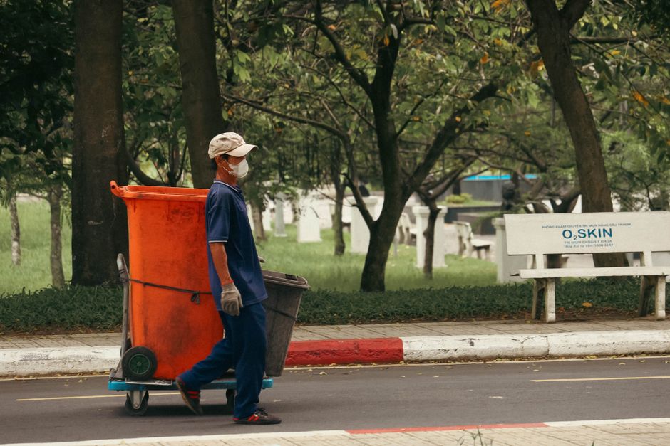 A waste collection truck fitted with a large rear hopper is actively engaged in rubbish removal, with a sanitation worker dressed in a yellow safety suit and helmet operating the lifting mechanism. The truck's exterior body is painted in a light grey colour with visible dirt and grime, indicating regular use. Several white and black garbage sacks, along with miscellaneous waste, are piled inside the open hopper, ready for disposal or transfer. The vehicle is positioned on an urban street, with some green-leaved trees and a modern building in the background, providing natural daylight that illuminates the scene. The surrounding environment suggests an area designated for waste collection, possibly on a municipal or private site. This scene represents the routine activity undertaken by waste management companies, such as rubbishremovalkingstonuponthames.com, which specialise in private waste disposal services. The truck's design and waste materials emphasize the importance of efficient rubbish removal and alternative waste handling outside traditional council services, supporting a professional approach to rubbish collection and debris clearance.