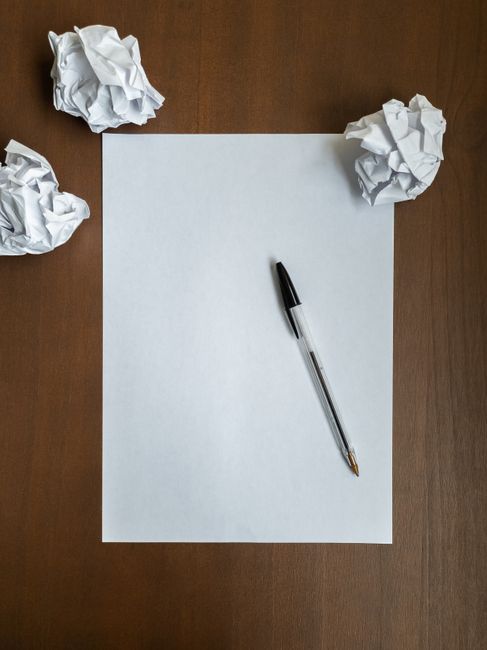 A blank white sheet of paper is centrally positioned on a wooden surface, with a black and transparent ballpoint pen resting diagonally across the lower right corner of the paper. Surrounding the paper are three crumpled sheets of white paper placed at irregular angles—one at the top left, one at the top right, and one at the bottom left—each appearing wrinkled and discarded. The wooden surface has a smooth, polished finish with a warm, brown tone, providing a clear contrast to the white paper and crumpled sheets. The lighting in the scene is even, highlighting the textures of the crumpled paper and the clean, flat surface of the large sheet, creating a natural environment that evokes ideas of planning or idea refinement, common in the context of rubbish removal planning or waste management strategies, with the scene subtly suggesting the process of eliminating unnecessary or unwanted materials. The overall composition is neat and professional, suitable for an article focused on avoiding common mistakes in private waste collection or alternative rubbish clearance methods.