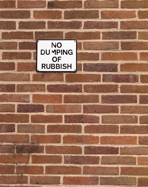 A rectangular white metal sign with black text that reads 'NO DUMPING OF RUBBISH' is mounted on a red brick wall. The bricks are laid in a horizontal pattern with varying shades of reddish-brown and some darker bricks, giving a textured appearance. The sign is positioned slightly above the center of the image and is attached to the wall with four visible screws, one at each corner. The wall appears clean and well-maintained, with no visible debris or clutter nearby, emphasizing a clear message against illegal rubbish dumping, consistent with the theme of responsible waste management and rubbish removal services. Natural lighting casts subtle shadows beneath the sign, highlighting the bricks’ rough surface and the sign’s smooth finish, creating a neutral yet official tone suitable for informing about proper waste disposal practices.