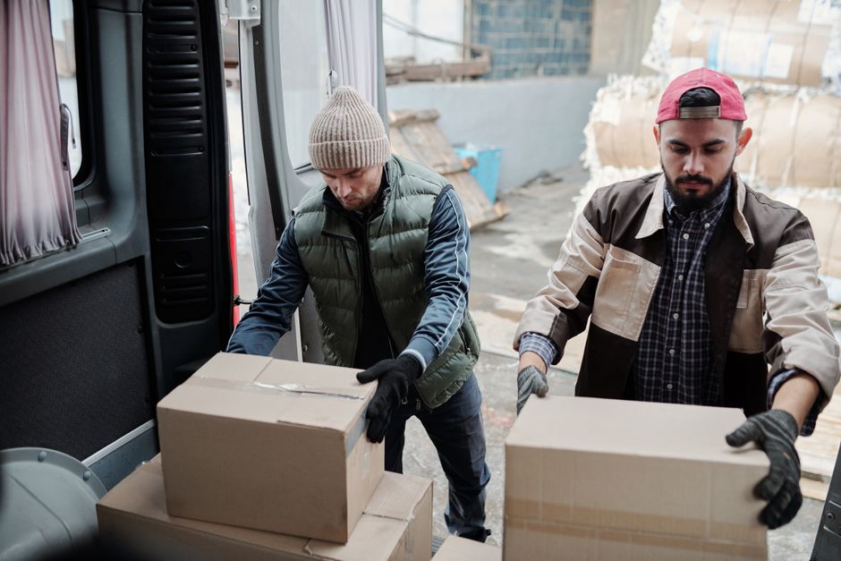 Two men wearing casual work attire and gloves are loading a cardboard box into the back of a grey commercial van with an open side door. The man on the left is wearing a beige knit beanie, a black puffer vest over a navy long-sleeve shirt, and dark pants, while the man on the right is sporting a red cap worn backwards, a multi-tone beige and black jacket, and a checked shirt underneath. They are standing on a paved outdoor area near a warehouse or storage facility, with some cardboard and other materials visible in the background. The interior of the van shows a dark plastic panel and a window with a partially drawn curtain. The scene suggests an activity related to waste disposal or rubbish collection, fitting with a professional rubbish removal service such as offered by Kingston-based companies that provide independent collection and private waste handling, as indicated on the webpage about permit requirements for van loading and rubbish removal in Kingston.