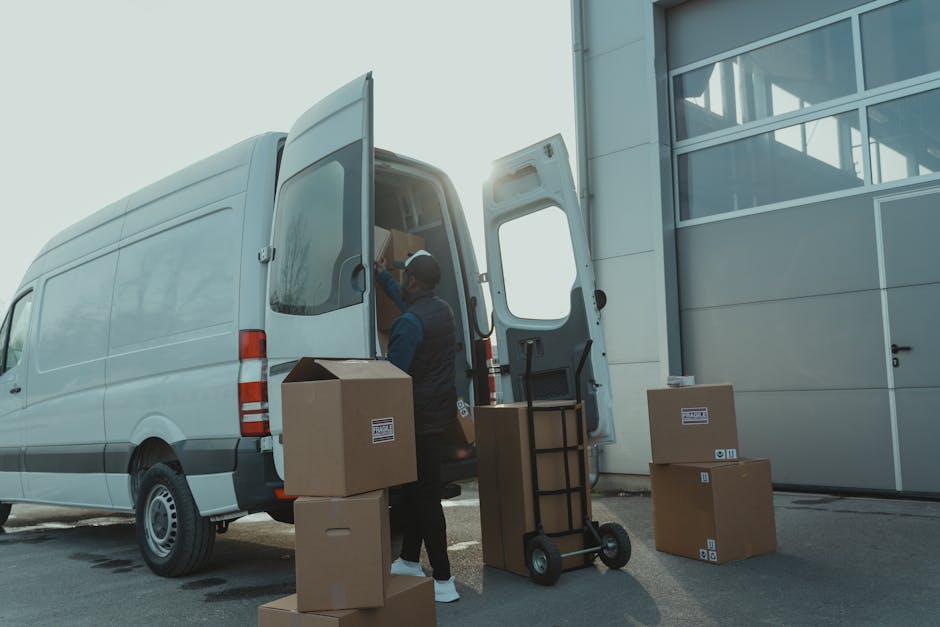 A person wearing a blue long-sleeve shirt and a black puffer vest is standing in front of an open delivery van, holding a clipboard in their left hand while their right hand is extended towards the clipboard. The van’s rear doors are open, revealing stacks of cardboard boxes with various labels and barcodes, some partially visible and arranged on shelving units inside the vehicle. The boxes vary in size and are made of brown corrugated cardboard, with some displaying white labels or packing tape. A large cardboard box is positioned in the foreground, partially obscuring the lower part of the individual’s body. The scene appears to occur in an outdoor setting, with the van parked on a paved surface. The environment suggests a logistics or waste management context, where packaging or rubbish is being organized for collection or disposal, reflecting the activities related to private waste handling and rubbish removal services provided by companies like rubbishremovalkingstonuponthames.com.