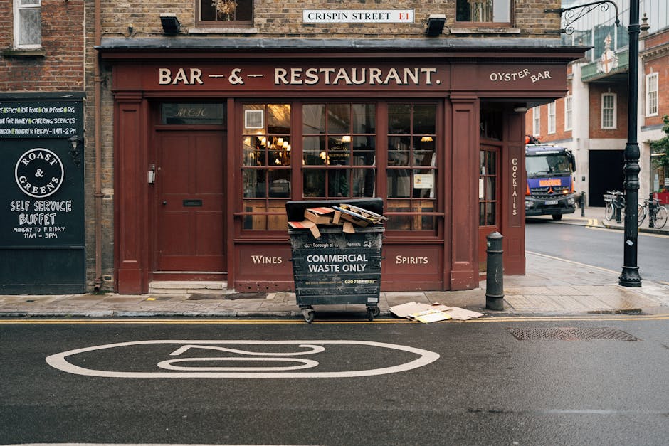 A street-facing view of a small restaurant or pub with a dark red-brown wooden exterior, featuring large glass windows revealing warm interior lighting. To the left, a black signboard displays menu information and opening hours. In front of the establishment, a large black wheelie bin labeled 'COMMERCIAL WASTE ONLY' is placed on the pavement, overflowing with flattened cardboard boxes and paper packaging, some spilling onto the ground. The bin is positioned close to a short black bollard and a drain cover on the wet asphalt road marked with a white speed limit sign. An older truck with a blue cab and orange hazard lights is visible parked further down the street, with bicycles secured to a bike rack near the corner. The surrounding environment indicates an urban area with brick buildings and a lamppost. THIS scene reflects a typical setting where private waste collection, such as commercial rubbish removal, might be managed by services like rubbishremovalkingstonuponthames.com, rather than relying solely on municipal refuse services.