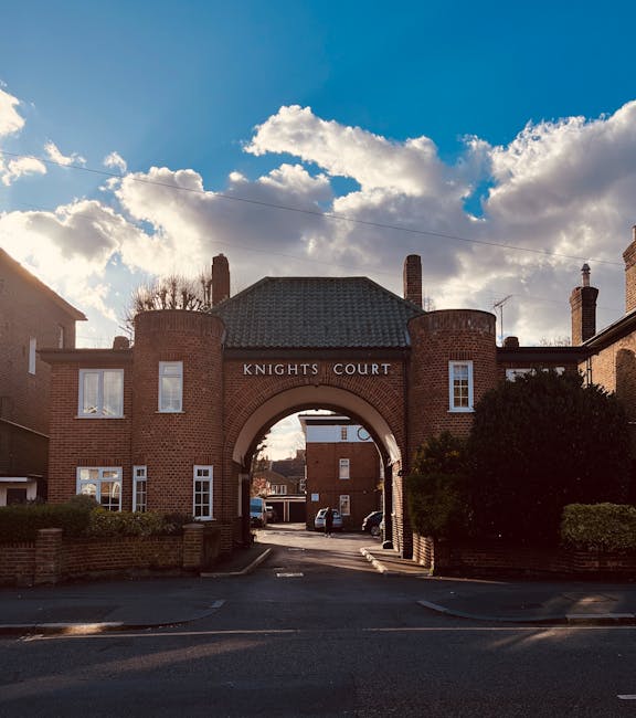 A brick archway entrance labeled 'Knights Court' is centrally positioned in the image, featuring a curved arch supported by two round brick towers on either side, with small rectangular windows. The brickwork is a reddish-brown tone with a textured surface. Above the arch, a gabled roof with dark shingles extends across, with a chimney visible on each side. Behind the arch, a narrow street or driveway leads into a residential area, where several parked cars are partially visible. On the left and right sides of the arch, low brick walls topped with neatly trimmed bushes create a boundary along the pavement. The scene is illuminated by natural daylight, with a sky filled with scattered cumulus clouds and patches of blue, casting a bright but diffused light over the scene. This setting may relate to private residential or estate entrances, similar in context to independent waste collection and on-site clearance scenarios, as suggested by [COMPANY_NAME]'s rubbish removal services in Kingston upon Thames.