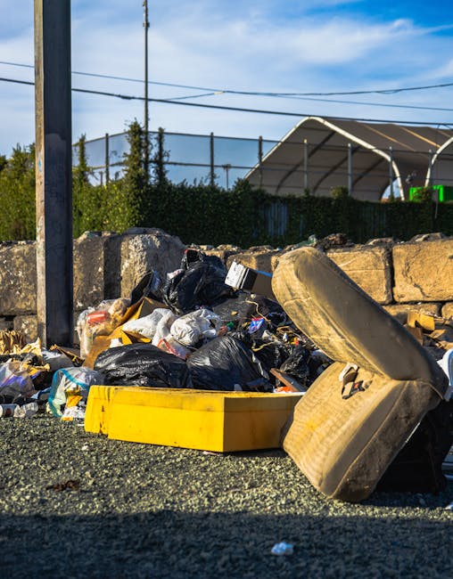 A large pile of mixed refuse and rubbish, including black plastic trash bags, a yellow plastic storage container, and a heavily soiled beige car tire, is situated on a gravel surface in an outdoor setting. The rubbish appears to consist of various household waste materials, with some bags partially torn open, revealing contents inside. In the background, there is a weathered stone wall composed of irregularly shaped stones, behind which green foliage and a tall metal fence are visible, along with a tennis or sports court lighted with bright green illumination. Overhead, electrical wires cross the clear partly cloudy sky, suggesting an urban or suburban environment. The scene hints at a private rubbish collection or on-site waste removal, possibly coordinated by a local provider such as rubbishremovalkingstonuponthames.com, focusing on non-municipal disposal of household or business waste materials in this area.