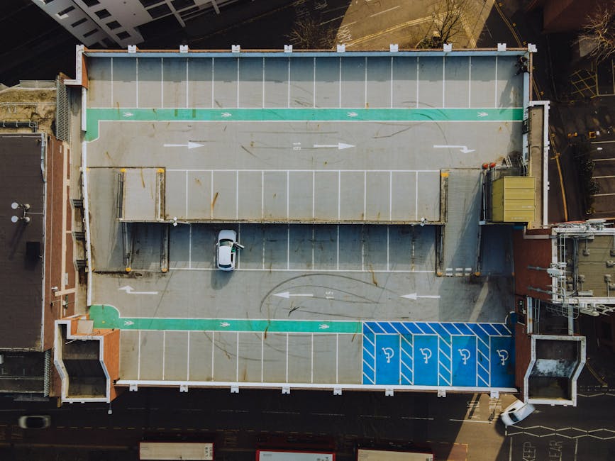 An aerial view of a rooftop parking area on a multi-storey building shows marked parking spaces, including designated accessible parking bays with blue pavement markings in the lower right corner. The parking lot surface is made of concrete with visible wear and dirt stains, and features white directional arrows and parking lines. In the center, a small white car is parked within a space, adjacent to a raised structure with a flat, rust-stained metal roof, supported by vertical metal beams. The rooftop is bordered by a parapet wall with a green strip along the top edge, serving as a safety barrier. Surrounding the building, parts of the cityscape including brick and concrete buildings and a street with vehicles and trees can be seen, with a mix of shaded and sunlit areas creating a dynamic lighting environment. The scene is indicative of a private parking or service area that could be associated with building maintenance or waste removal operations, situated within an urban context.