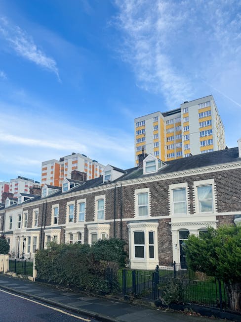 A row of traditional brick terraced houses with white window frames and decorative detailing along the upper facades, situated on a paved street with small front gardens enclosed by black metal fences. The gardens are planted with bushes and small trees, with some greenery visible close to the pavement. In the background, modern high-rise apartment buildings with yellow and white exteriors rise against a partly cloudy blue sky, highlighting a contrast between old and new urban architecture. The scene is well-lit, likely during daytime, with natural light illuminating the buildings and casting soft shadows. This setting reflects a typical residential area where alternative waste handling services, such as private rubbish removal, might be needed for local property management or renovation waste clearance, supported by companies like rubbishremovalkingstonuponthames.com.