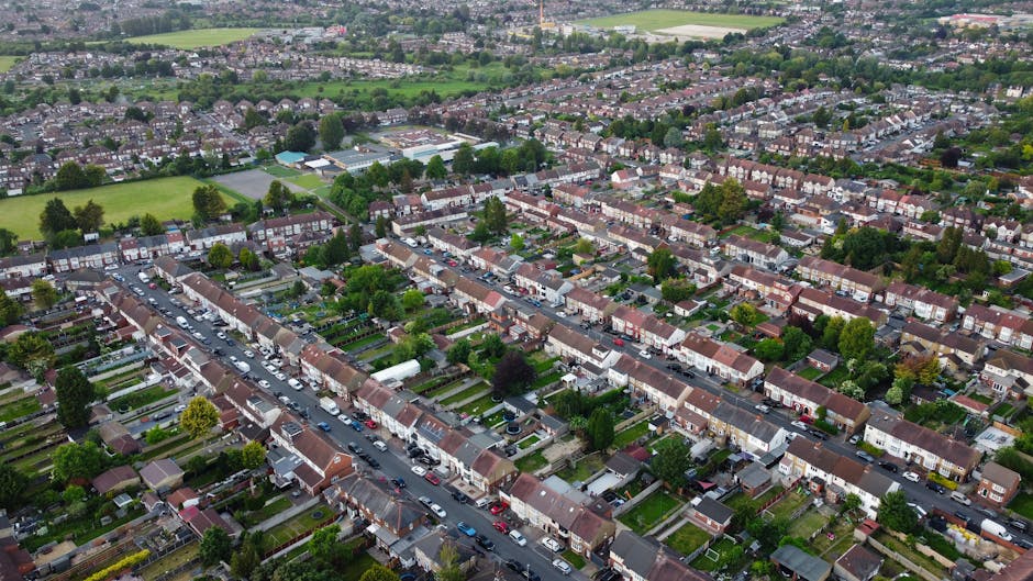 An aerial view of a residential area with rows of terraced houses featuring brick facades and tiled roofs, lining a straight street with parked cars along both sides. The front gardens vary in size, with some having neatly maintained lawns, flower beds, and small trees. In the background, there are green open spaces and sports fields, bordered by additional housing and trees, under a cloudy sky with diffused light. The scene captures a typical suburban neighbourhood likely within Kingston upon Thames, illustrating the density and character of local housing, and providing context for private waste collection or rubbish removal services by companies like rubbishremovalkingstonuponthames.com.