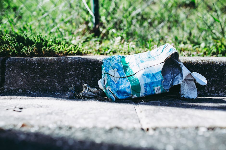 A crumpled plastic and paper bag lying on a concrete pavement next to a curb, partially filled with trash and debris, with rough textures and faded colors of blue, white, and green visible on the bag. The bag is situated close to the edge of the pavement, near a grassy area with lush green grass and small plants in the background, indicating an outdoor urban environment. Soft, natural lighting highlights the details of the bag and the surrounding concrete surface, which has small cracks and weathered patches. The scene suggests a discarded waste item awaiting collection, typical of unscheduled rubbish or private waste disposal, with no visible signs of active rubbish removal in progress. The setting reflects an outdoor area where rubbish is often left temporarily, emphasizing the importance of proper waste management and timely rubbish removal services, such as those provided by independent waste collection operators like rubbishremovalkingstonuponthames.com.
