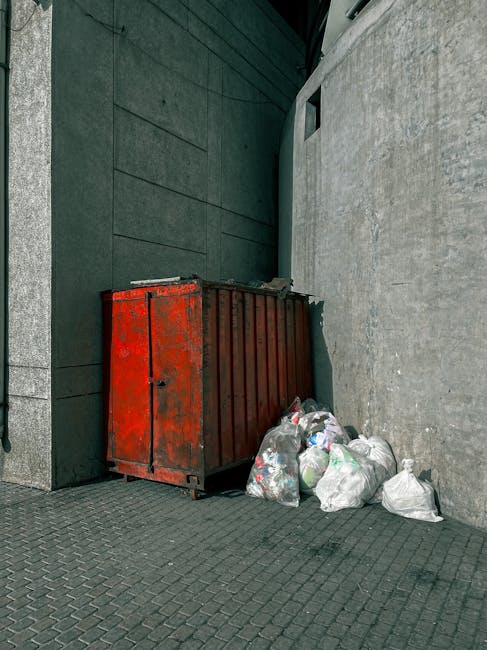A crumpled plastic and paper bag lying on a concrete pavement next to a curb, partially filled with trash and debris, with rough textures and faded colors of blue, white, and green visible on the bag. The bag is situated close to the edge of the pavement, near a grassy area with lush green grass and small plants in the background, indicating an outdoor urban environment. Soft, natural lighting highlights the details of the bag and the surrounding concrete surface, which has small cracks and weathered patches. The scene suggests a discarded waste item awaiting collection, typical of unscheduled rubbish or private waste disposal, with no visible signs of active rubbish removal in progress. The setting reflects an outdoor area where rubbish is often left temporarily, emphasizing the importance of proper waste management and timely rubbish removal services, such as those provided by independent waste collection operators like rubbishremovalkingstonuponthames.com.